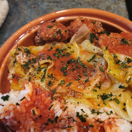 A plated serving of tender cooked cabbage, large meatballs, and rich tomato sauce served over a bed of white rice and garnished with fresh herbs.