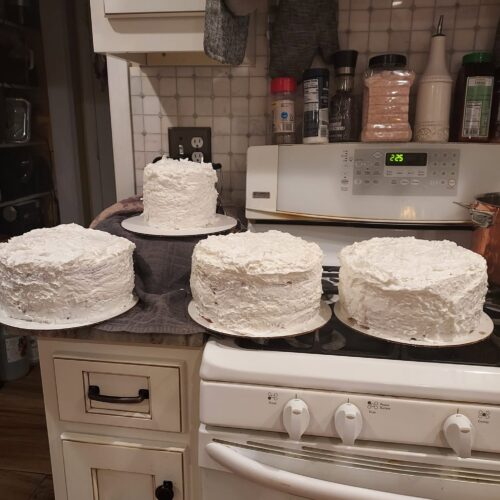 Four rustic, freshly frosted white coconut cakes sitting on a kitchen stove and counter.