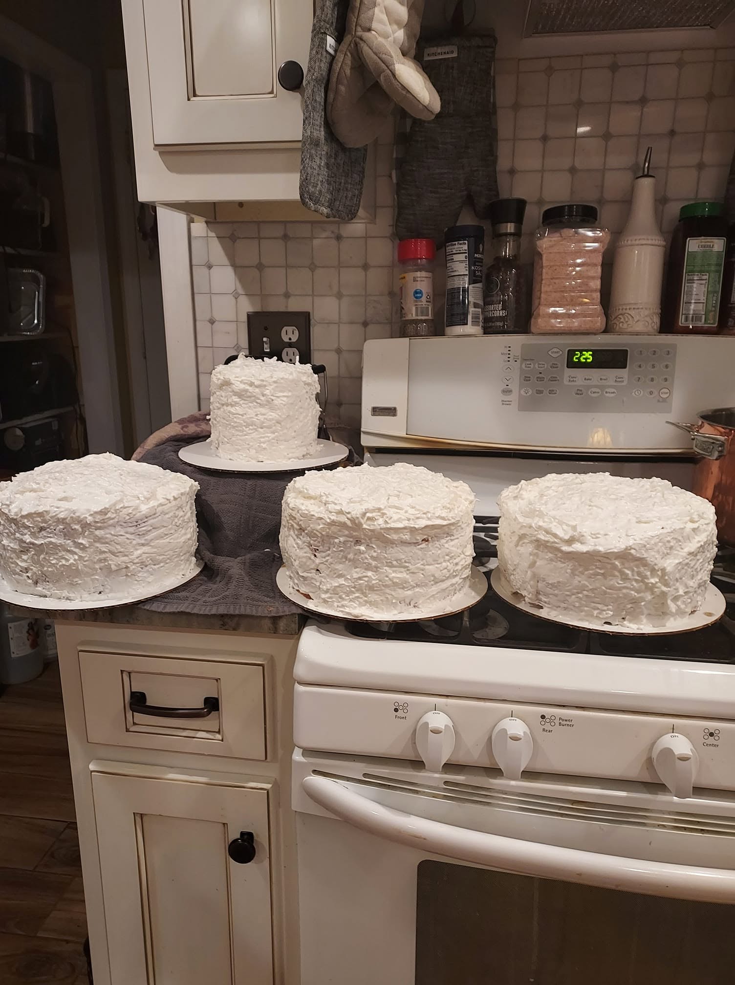 Four rustic, freshly frosted white coconut cakes sitting on a kitchen stove and counter.