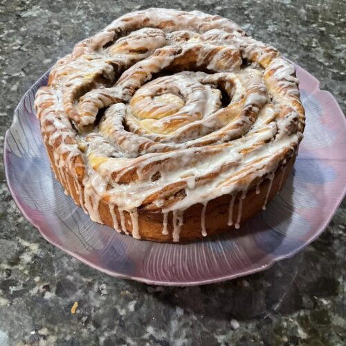 A freshly baked, giant cinnamon roll covered in a sweet vanilla and almond glaze, served on a decorative purple plate.