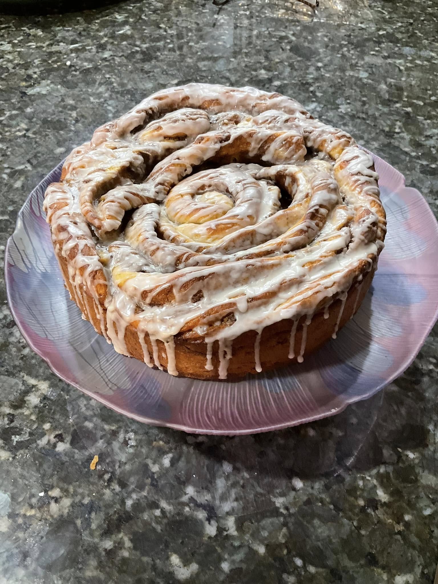 A freshly baked, giant cinnamon roll covered in a sweet vanilla and almond glaze, served on a decorative purple plate.