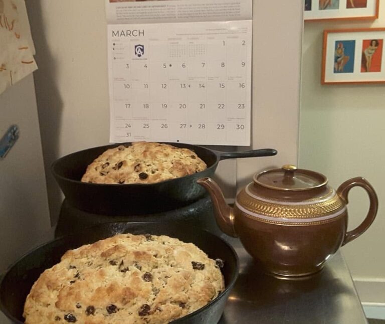 Two rustic loaves of freshly baked Irish soda bread sitting in black cast iron skillets next to a vintage brown teapot.