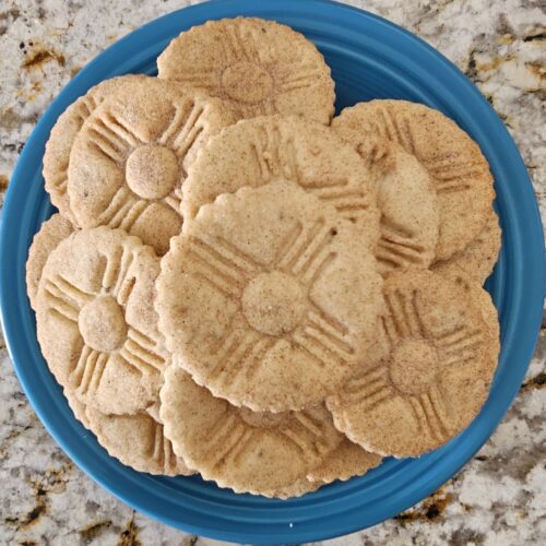A blue plate stacked with golden-brown New Mexico biscochitos stamped with the traditional sun symbol.