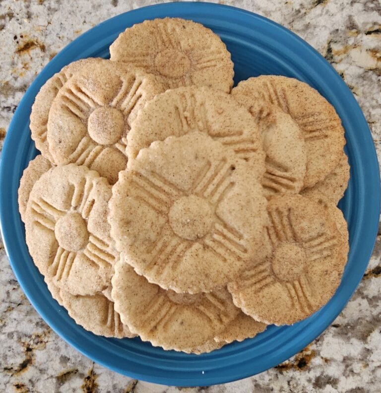 A blue plate stacked with golden-brown New Mexico biscochitos stamped with the traditional sun symbol.