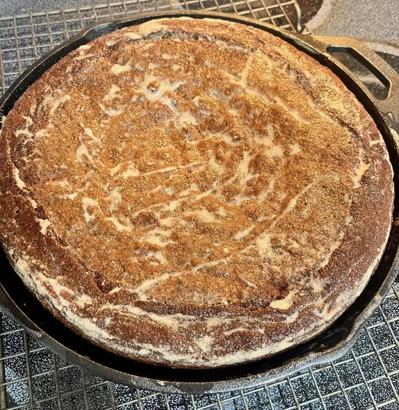 A beautiful, perfectly baked golden-brown banana bread resting in a round black cast iron skillet on top of a wire cooling rack.