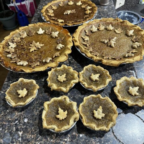 A countertop display of several freshly baked maple syrup pies and mini pies, all decorated with charming pastry maple leaf cutouts around the edges and centers.