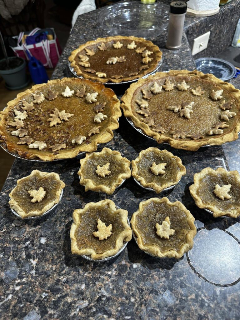 A countertop display of several freshly baked maple syrup pies and mini pies, all decorated with charming pastry maple leaf cutouts around the edges and centers.