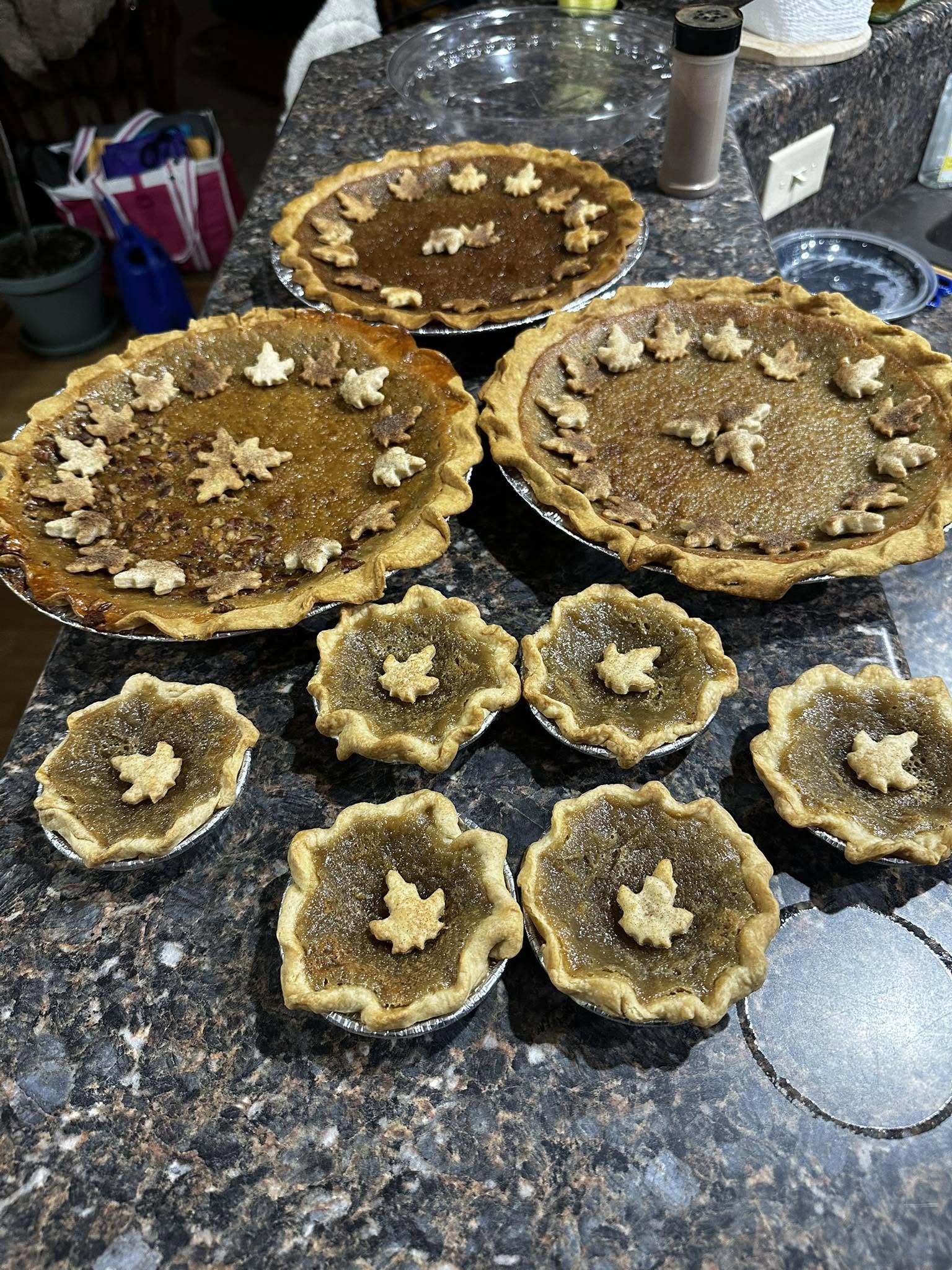 A countertop display of several freshly baked maple syrup pies and mini pies, all decorated with charming pastry maple leaf cutouts around the edges and centers.