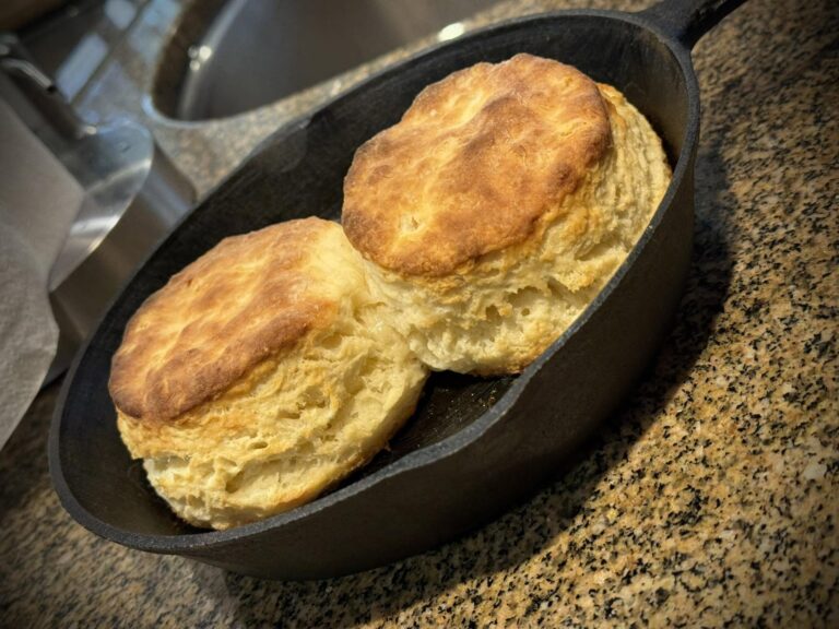 Two large, golden-brown flaky buttermilk biscuits baked in a small black cast iron skillet on a granite countertop.