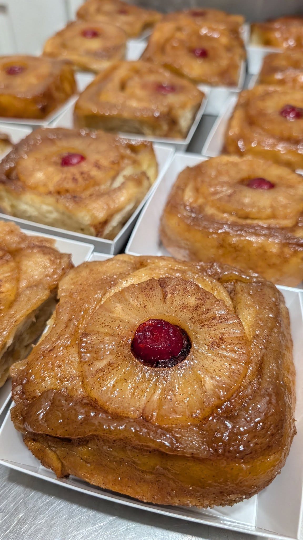 Beautifully glazed, square-shaped pineapple upside-down cinnamon rolls in individual white boxes, each topped with a caramelized pineapple ring and a bright red maraschino cherry.