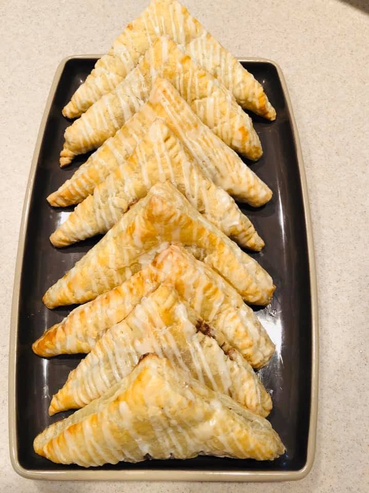 A dark rectangular platter stacked beautifully with eight golden-brown, triangle-shaped apple turnovers drizzled with a sweet vanilla glaze.