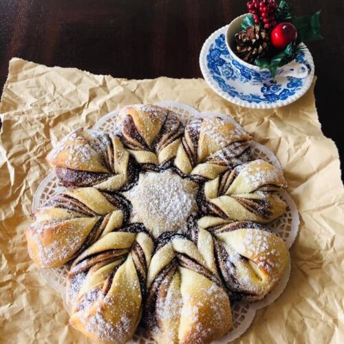 A beautiful, golden-brown baked Christmas star bread filled with chocolate hazelnut spread, heavily dusted with powdered sugar, and served on crinkled parchment paper next to a festive teacup.