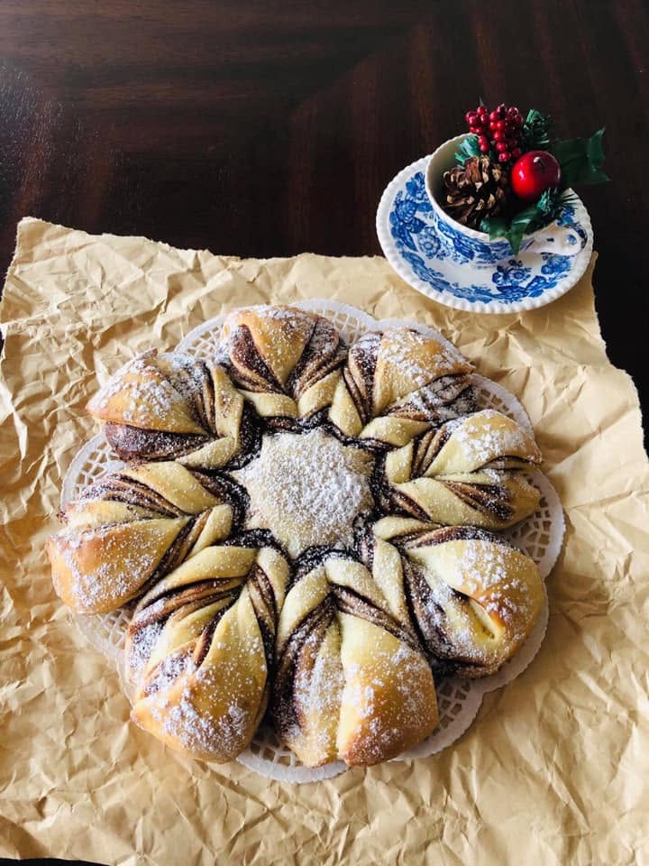 A beautiful, golden-brown baked Christmas star bread filled with chocolate hazelnut spread, heavily dusted with powdered sugar, and served on crinkled parchment paper next to a festive teacup.