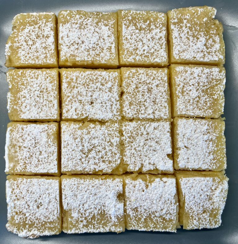 A white baking dish filled with gooey, bright yellow lemon bars heavily dusted with powdered sugar, with a silver fork lifting a freshly cut square slice.