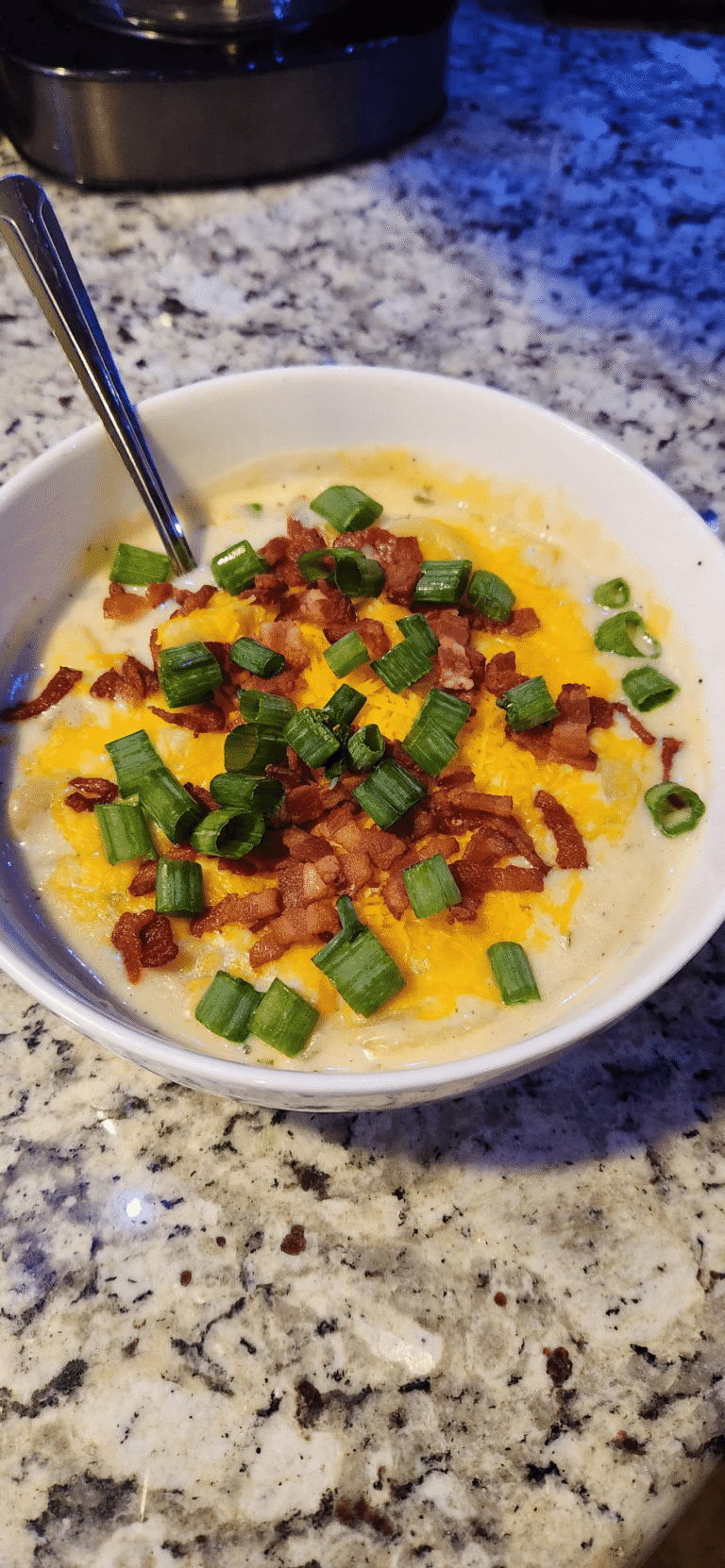 A large, rustic wooden bowl filled with rich, creamy potato soup topped with crisp bacon and green onions, with the full slow cooker pot resting on the counter in the background.