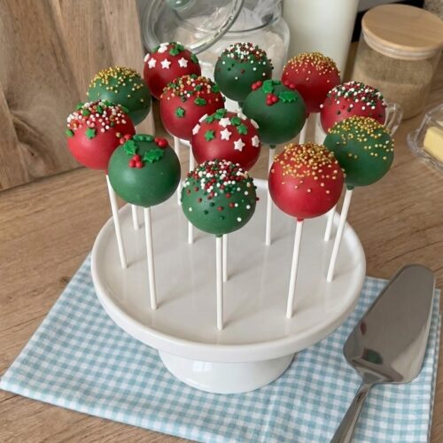 Red and green Christmas cake pops decorated with festive sprinkles and edible holly, displayed on white sticks in a cake pop stand.