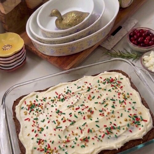 Homemade gingerbread cookie bars in a glass baking dish topped with thick cream cheese frosting and red, green, and gold Christmas sprinkles.