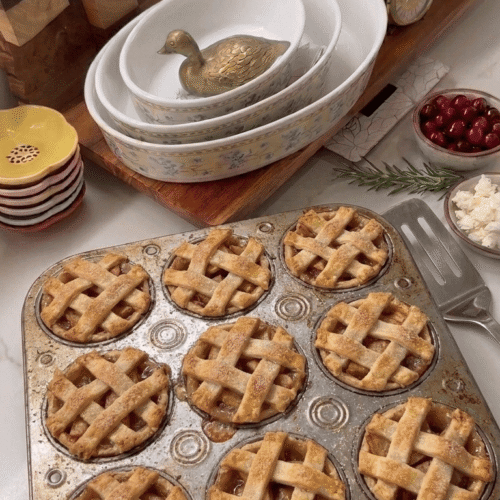 A freshly baked batch of mini apple pies with golden brown lattice crusts sitting in a rustic metal muffin tin on a kitchen counter.