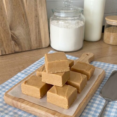 Thick squares of homemade 3-ingredient peanut butter fudge stacked on a wooden serving board, showing a smooth, creamy golden peanut butter texture.