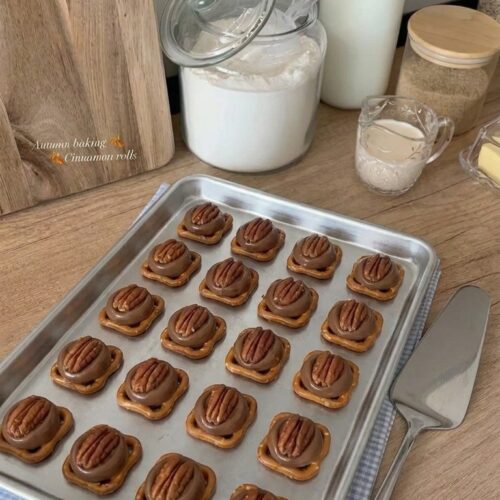 A silver baking sheet neatly lined with Rolo pretzel turtle bites featuring square pretzels, melted chocolate caramel candies, and pecan halves, resting on a kitchen counter.