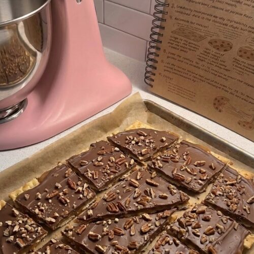 Homemade old-fashioned Pine Bark saltine toffee cut into pieces with melted chocolate and chopped pecans on a parchment-lined baking sheet.