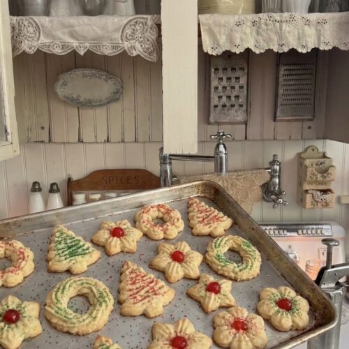 A vintage metal baking tray filled with freshly baked old-fashioned spritz butter cookies shaped like Christmas trees, wreaths, and stars, beautifully decorated with festive red and green sugar and candied cherries.