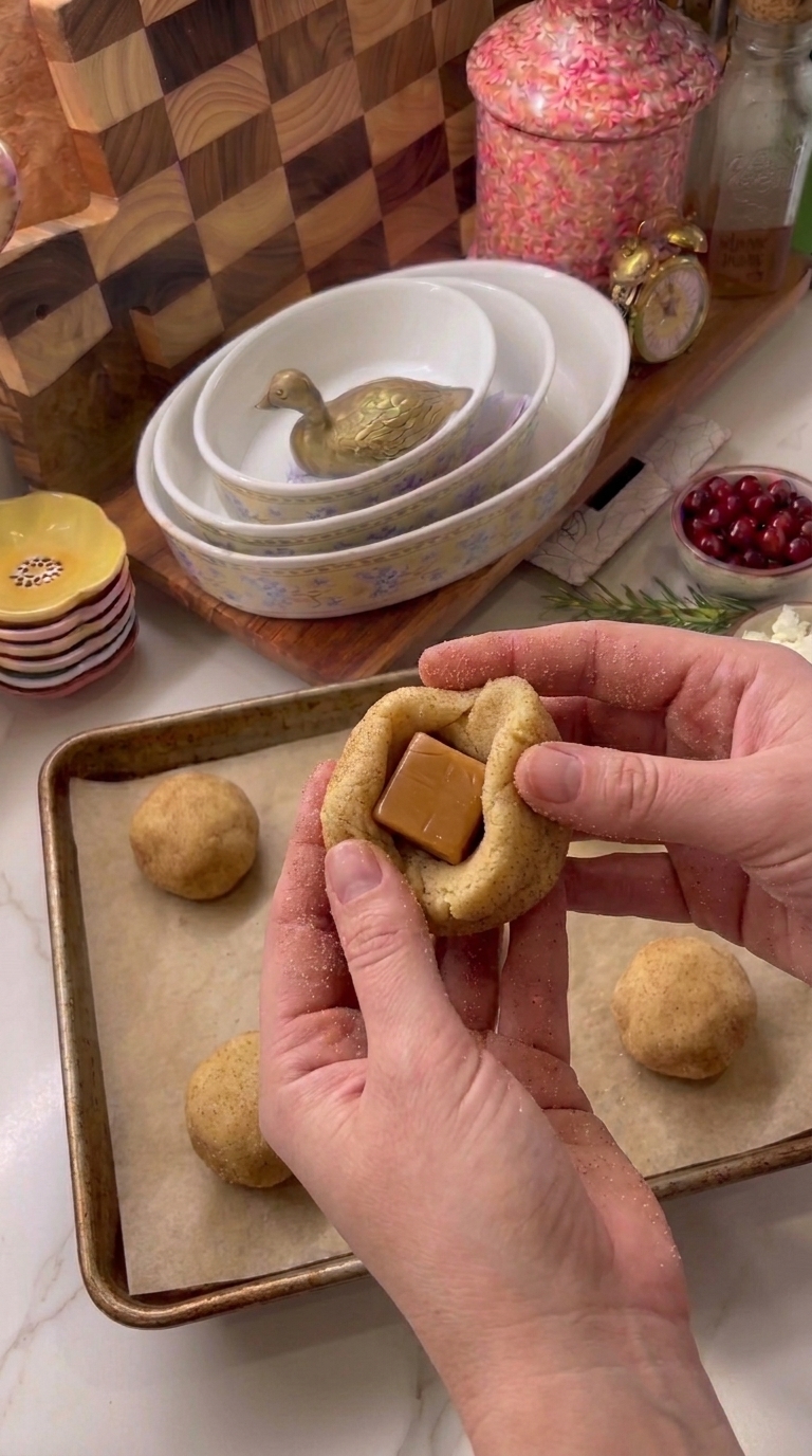 Hands wrapping soft snickerdoodle cookie dough around an unwrapped caramel candy before rolling it into a ball.