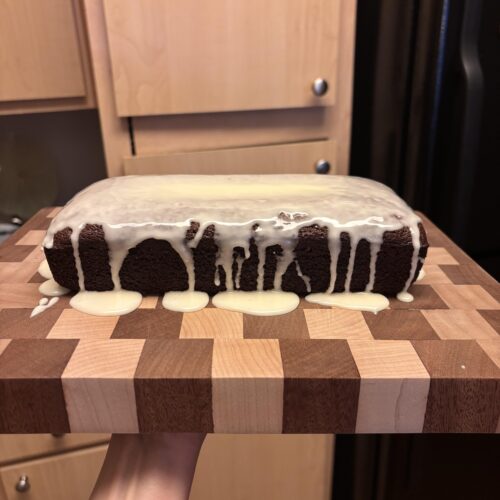 A sliced gingerbread loaf topped with a thick, sweet vanilla glaze, resting on a wooden cutting board with holiday spices scattered around.