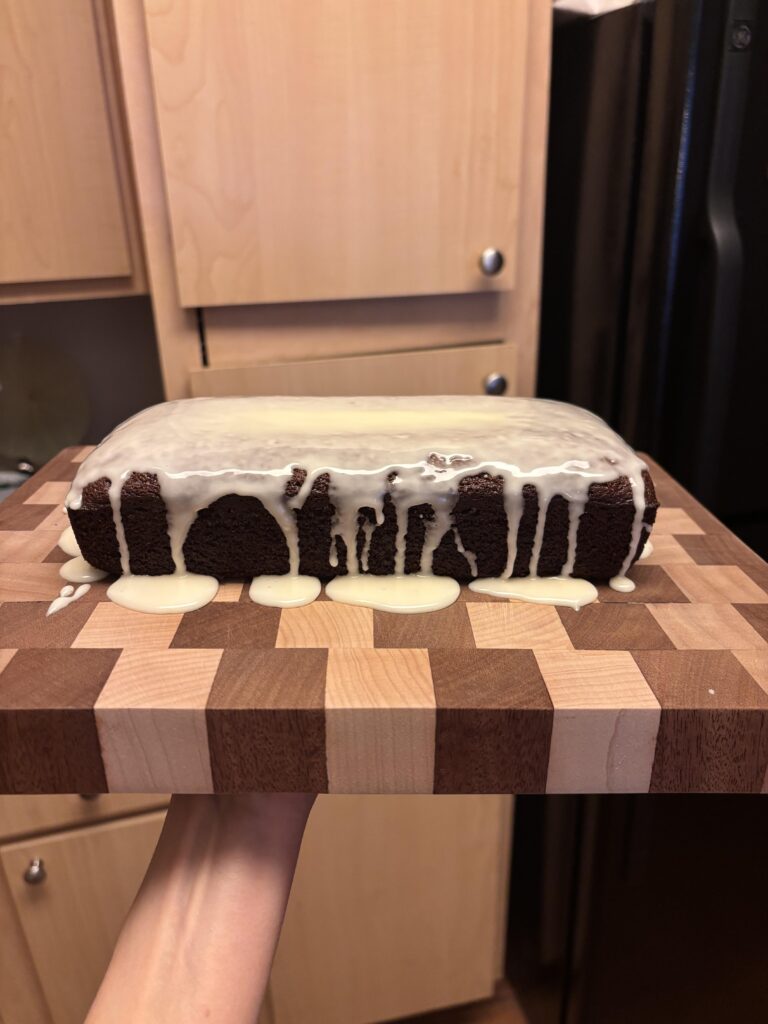 A sliced gingerbread loaf topped with a thick, sweet vanilla glaze, resting on a wooden cutting board with holiday spices scattered around.