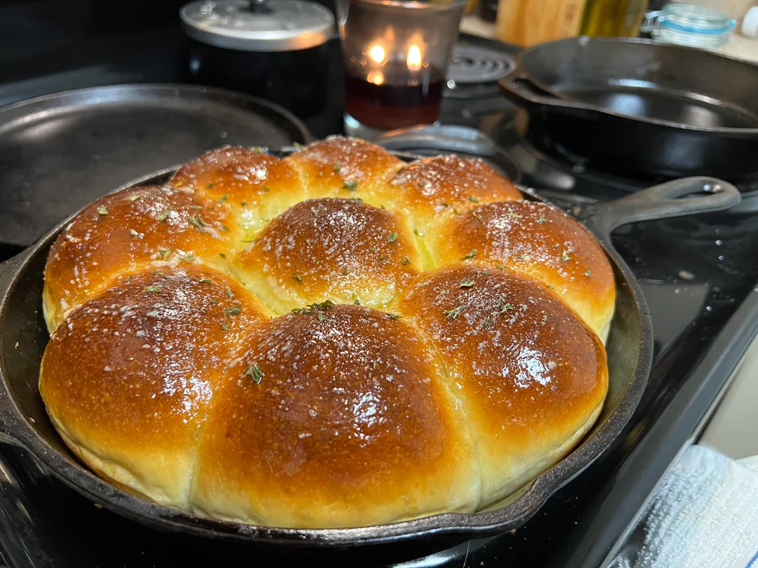 A well-seasoned cast iron skillet filled with eight golden brown, fluffy dinner rolls brushed with glistening herb butter.