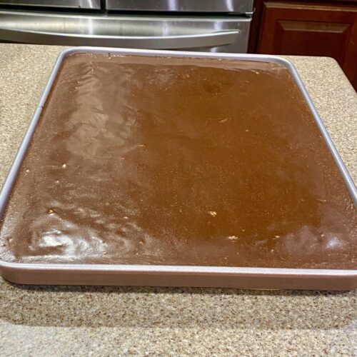A large, moist slice of chocolate fudge sheet cake dripping with warm poured chocolate icing and sprinkled with chopped pecans, being lifted from a baking pan.