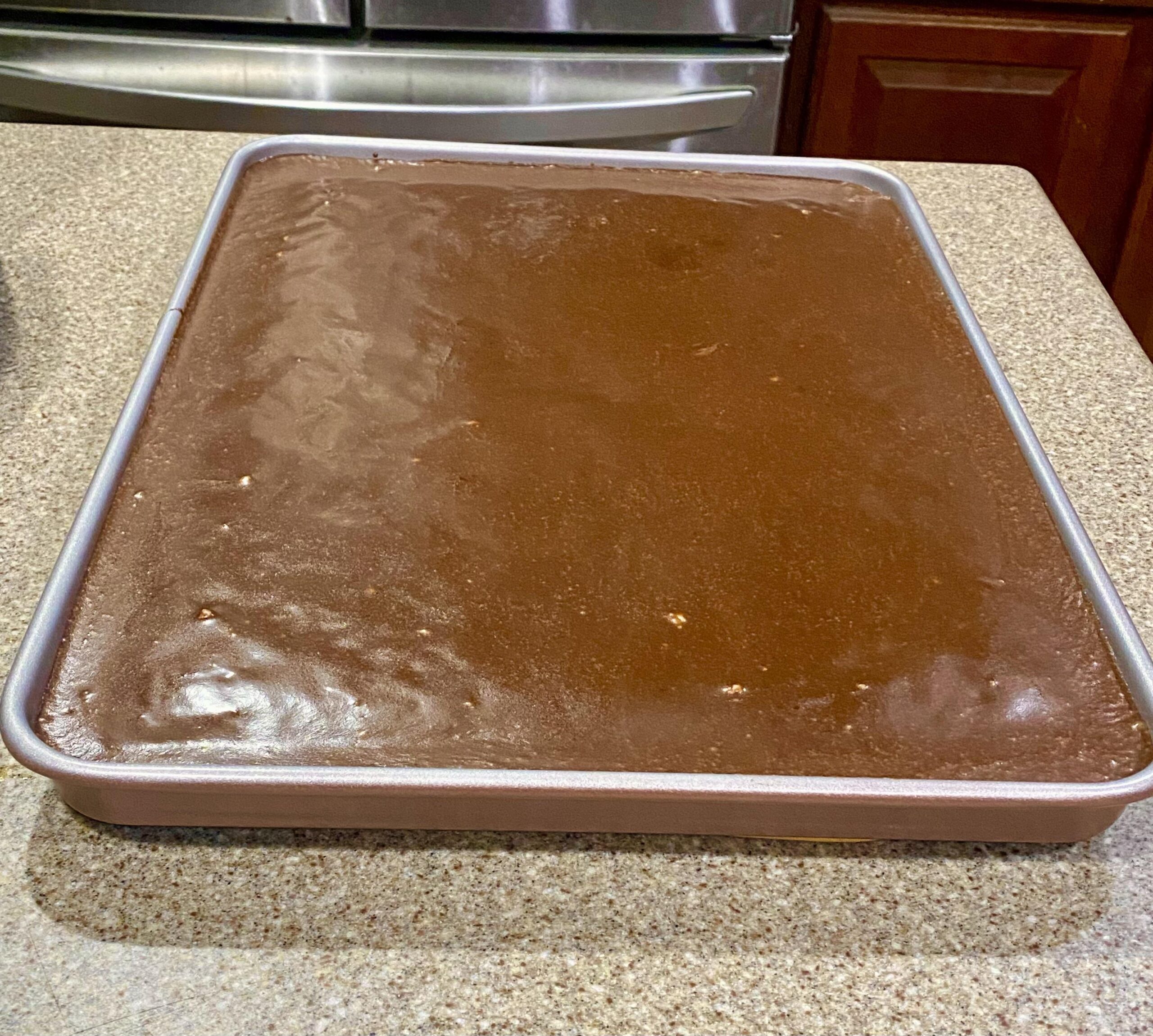 A large, moist slice of chocolate fudge sheet cake dripping with warm poured chocolate icing and sprinkled with chopped pecans, being lifted from a baking pan.