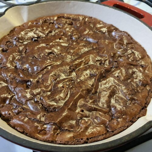 A large, round decadent fudgy brownie baked in a cast-iron skillet, featuring a perfect shiny, crinkly top crust, resting on a wooden cutting board.