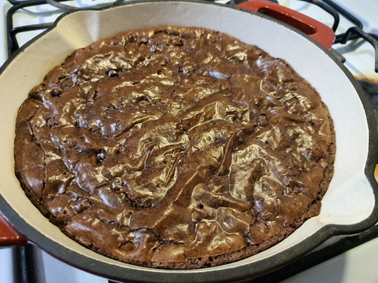 A large, round decadent fudgy brownie baked in a cast-iron skillet, featuring a perfect shiny, crinkly top crust, resting on a wooden cutting board.